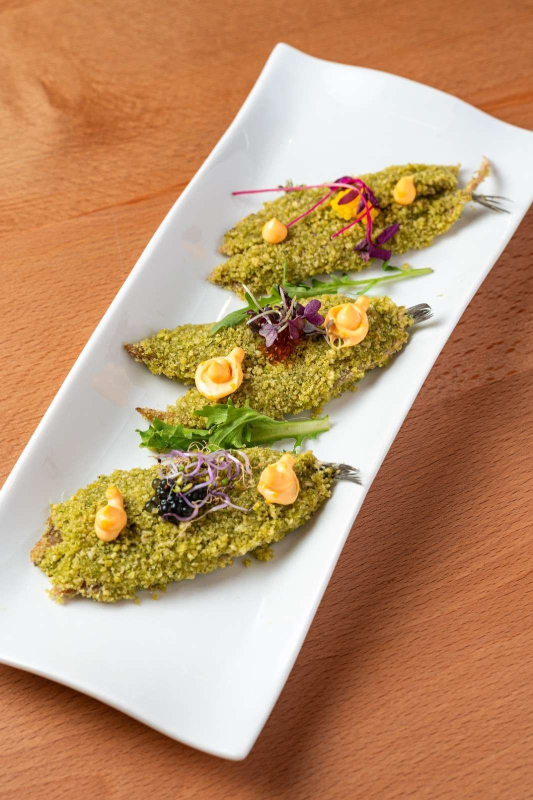 Cuatro aperitivos rebozados en hierbas verdes sobre un plato blanco, adornados con brotes morados y salsa amarilla.
