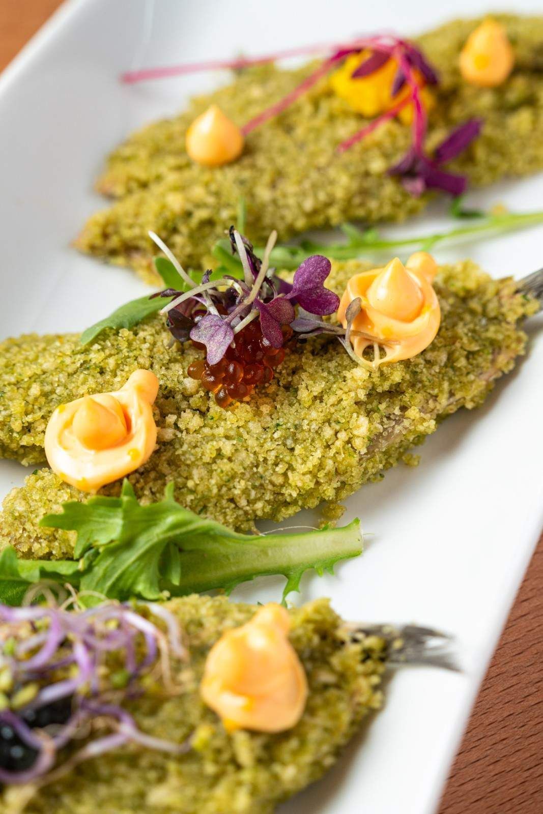 Filetes de pescado empanizados rebozados con hierbas verdes, adornados con verduras y salsa de naranja en un plato blanco.