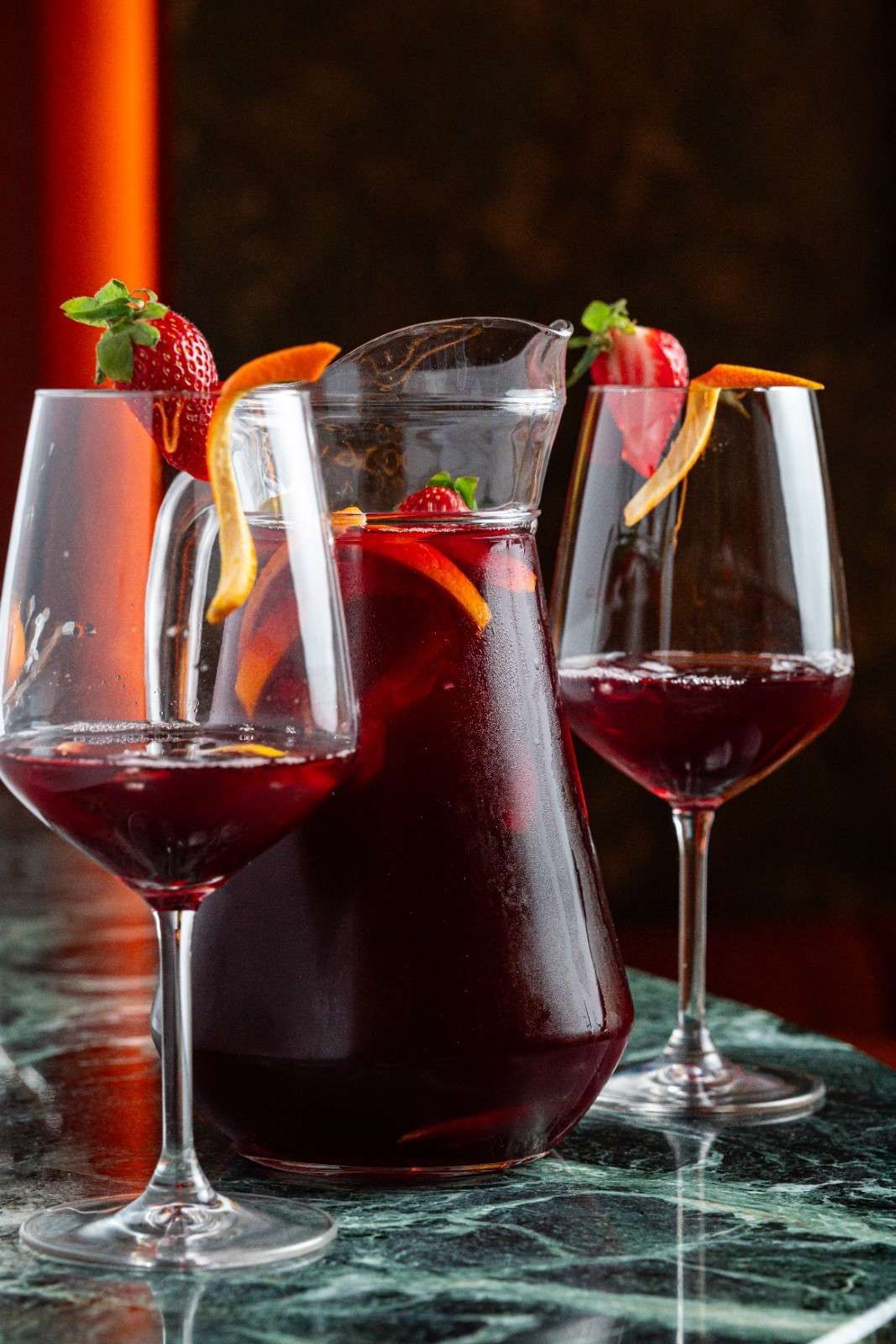 Tres copas de vino tinto y una jarra con fresas sobre una mesa de mármol.