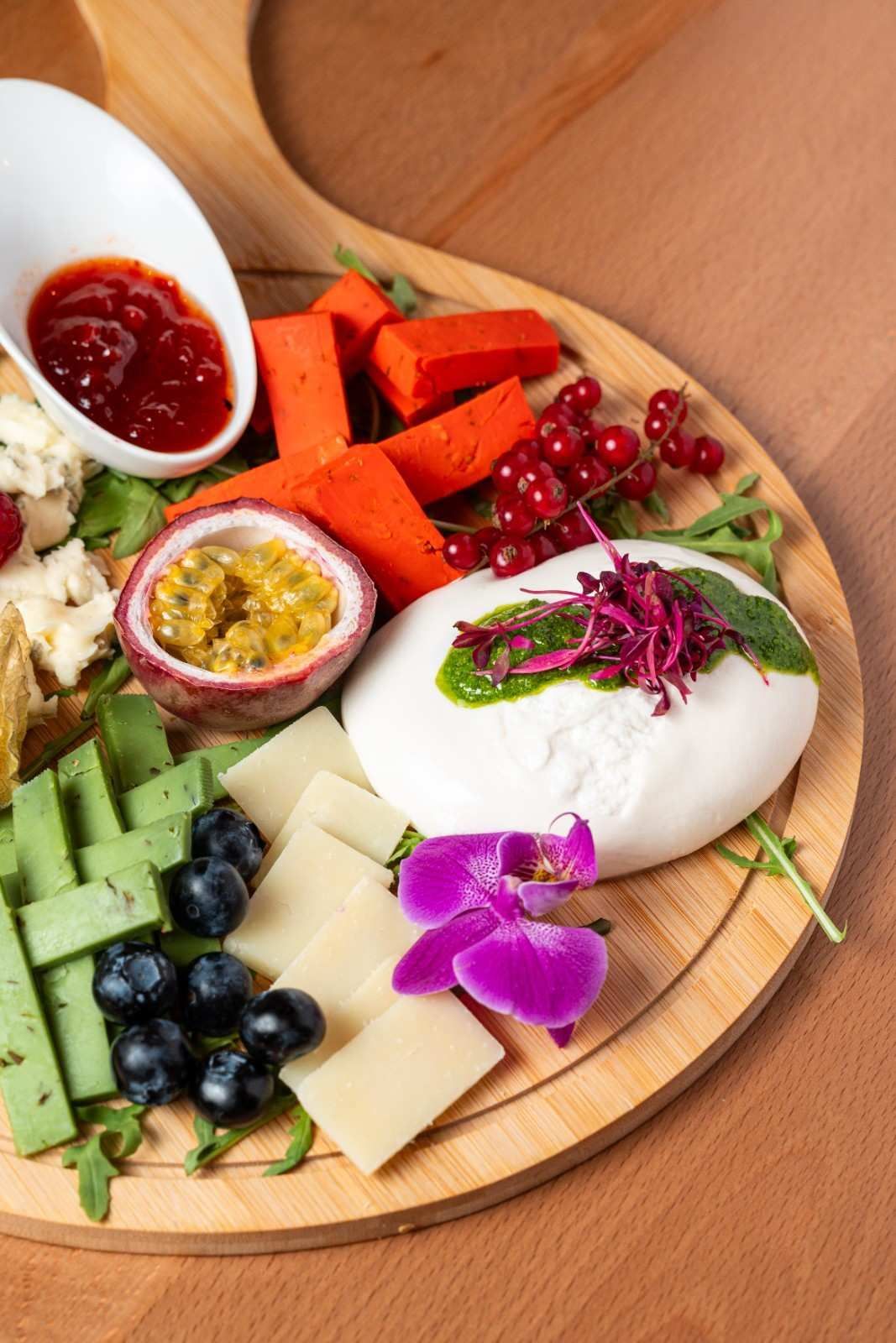 Tabla de quesos con fruta, mermelada, frutos secos, verduras y flores comestibles moradas en una bandeja de madera.