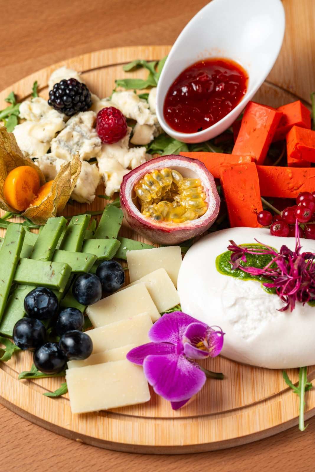 Tabla de embutidos con quesos, fruta, galletas saladas, mermeladas y flores comestibles en una bandeja de madera.