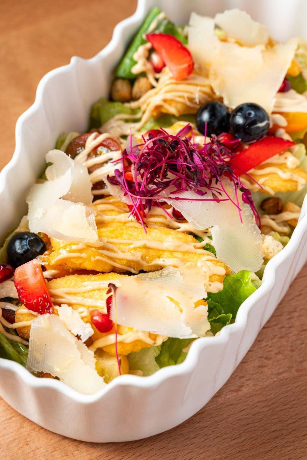 Ensalada colorida en un plato ovalado blanco con verduras, bayas y queso rallado sobre una superficie de madera.
