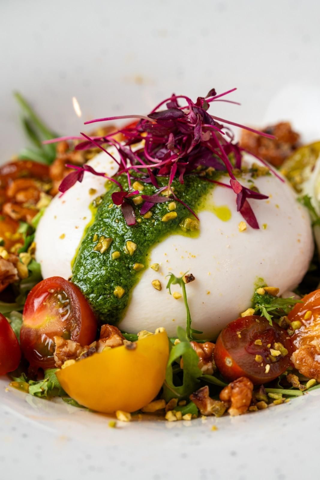 Burrata cubierta con pesto verde, tomates cherry y microbrotes morados en un tazón blanco.