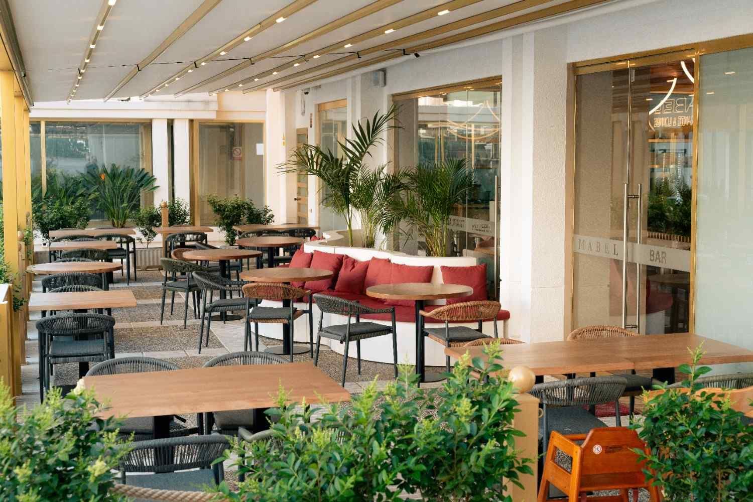 Terraza vacía de una cafetería al aire libre con mesas de madera, sillas negras, bancos rojos y plantas