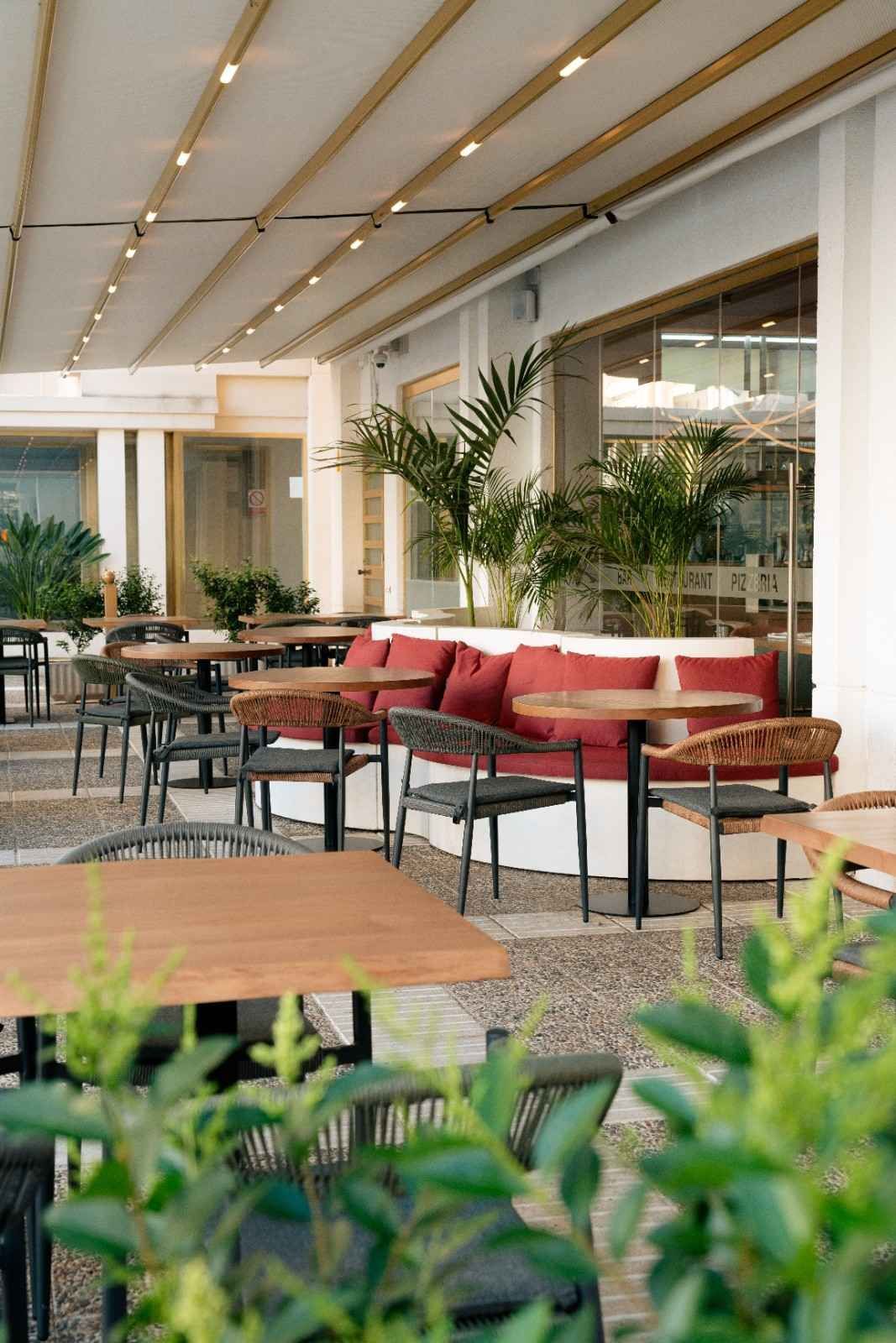 Terraza moderna con asientos tipo café, cojines rojos, plantas en macetas y un patio cubierto.