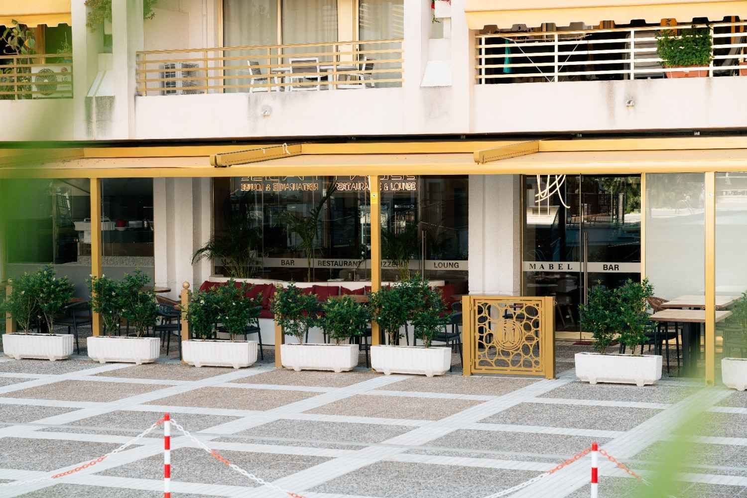 Cafetería al aire libre con plantas en macetas, mesas y pavimento a rayas, ubicada fuera de un edificio bajo un balcón.