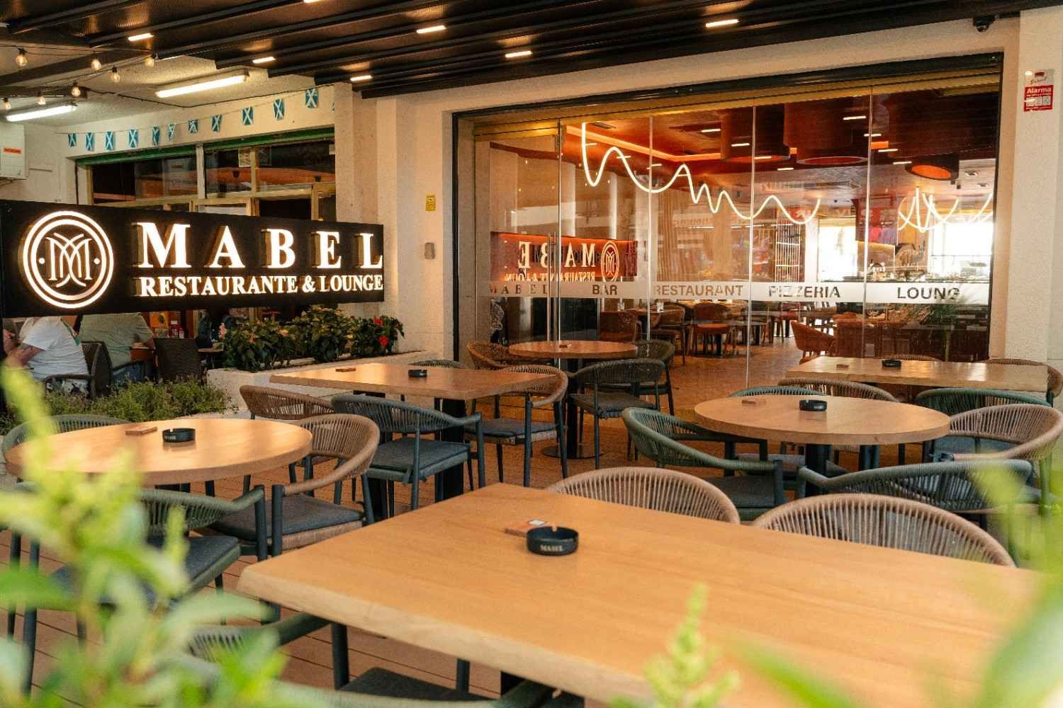 Patio del restaurante y salón Mabel con mesas de madera, sillas de mimbre e interior iluminado