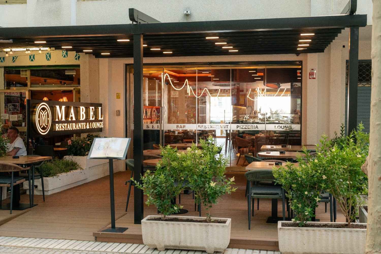 Entrada del restaurante que da a la calle, con mesas al aire libre, plantas verdes y un letrero que dice