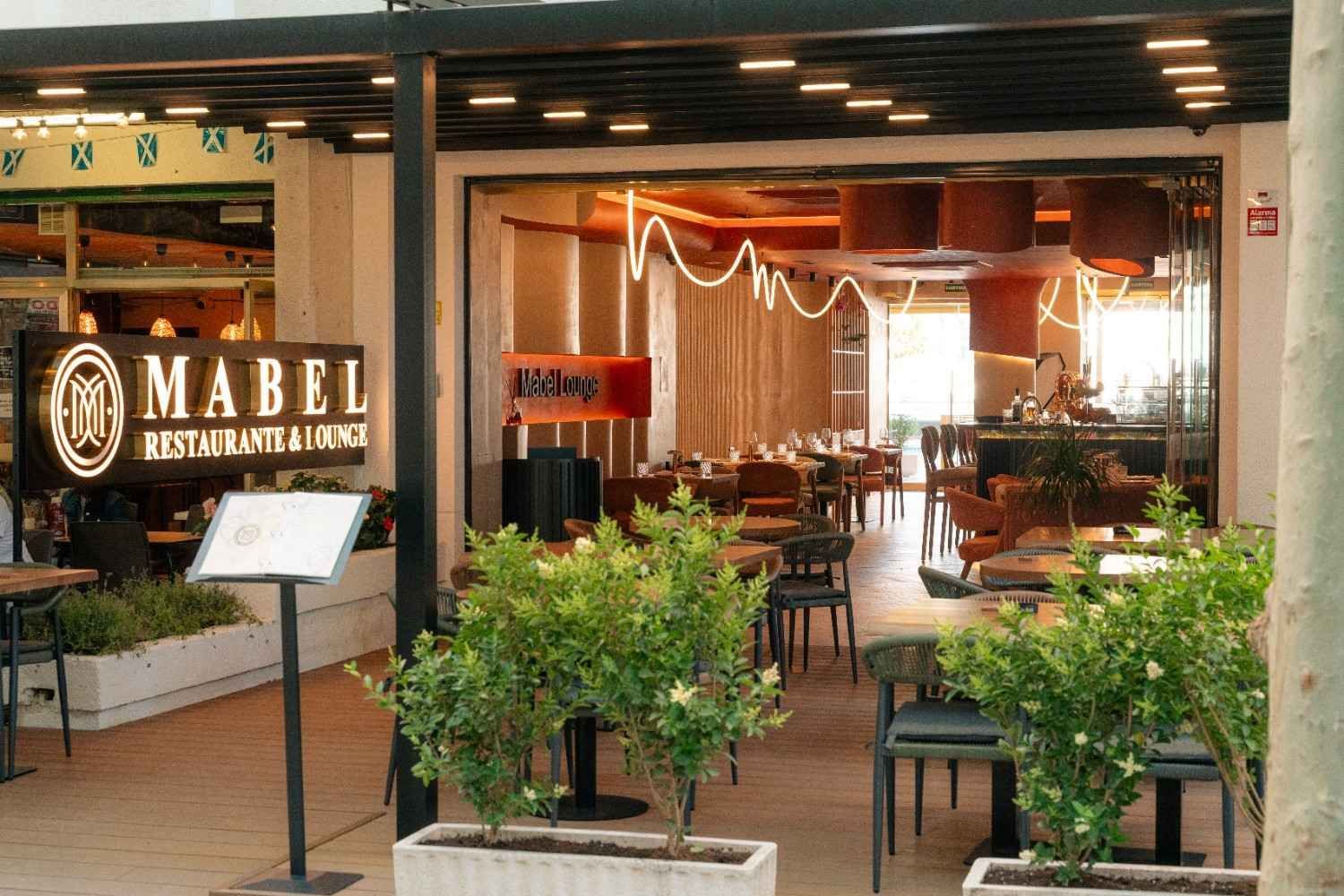 Entrada exterior al restaurante Mabel con plantas y zona de comedor con iluminación cálida en el interior.