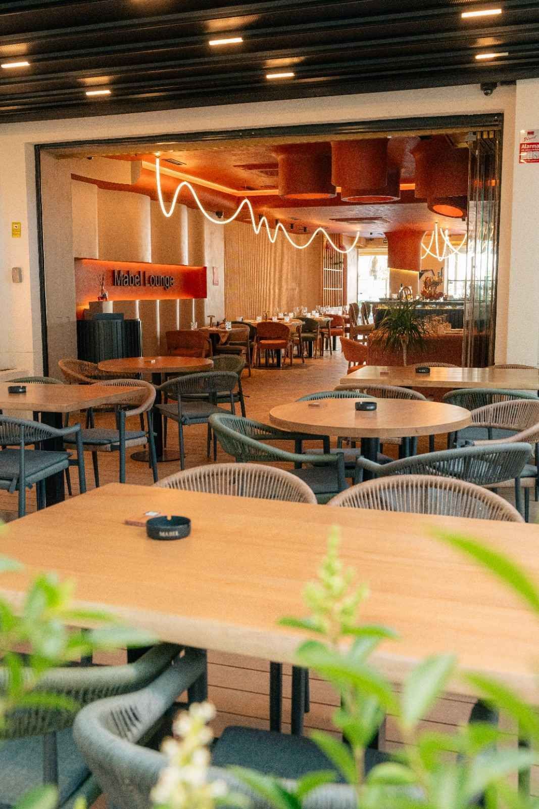 Cafetería vacía con mesas redondas y sillas de mimbre, un interior de madera cálida y guirnaldas de luces colgando del techo.