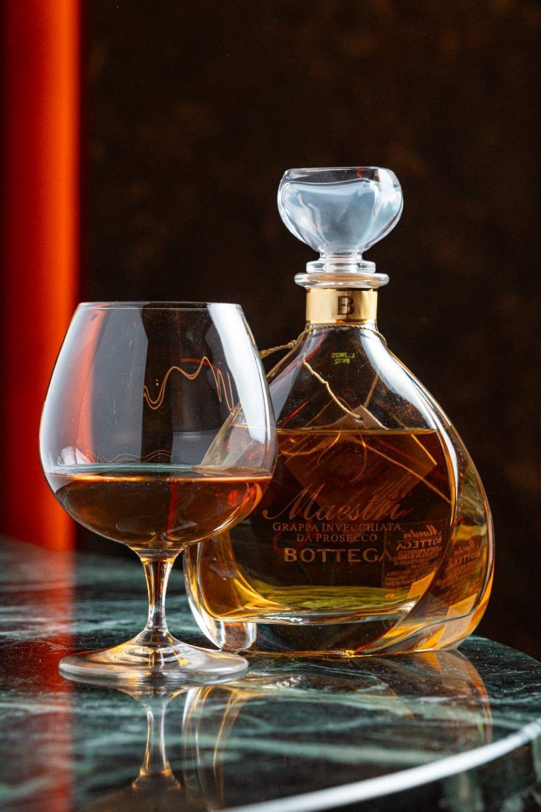 Un vaso de whisky y una jarra de cristal con licor ámbar sobre una mesa reflectante con fondo oscuro.
