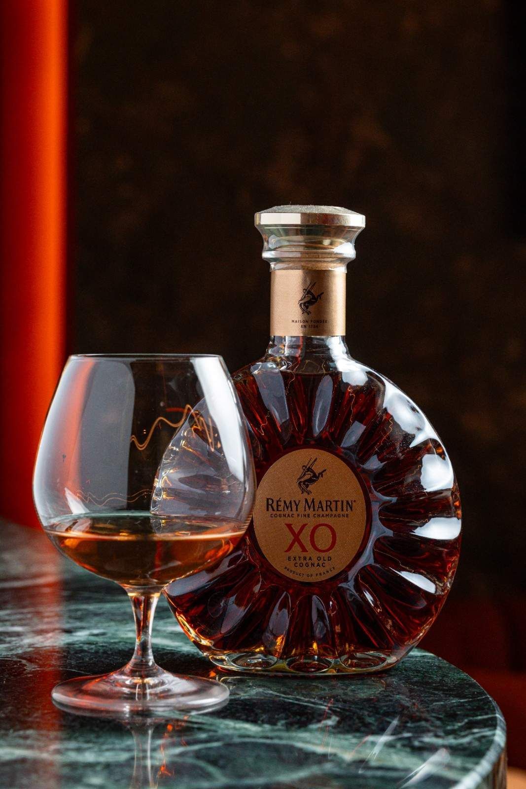 Una copa de coñac junto a una botella redonda de XO sobre una mesa de mármol oscuro con iluminación ámbar cálida.