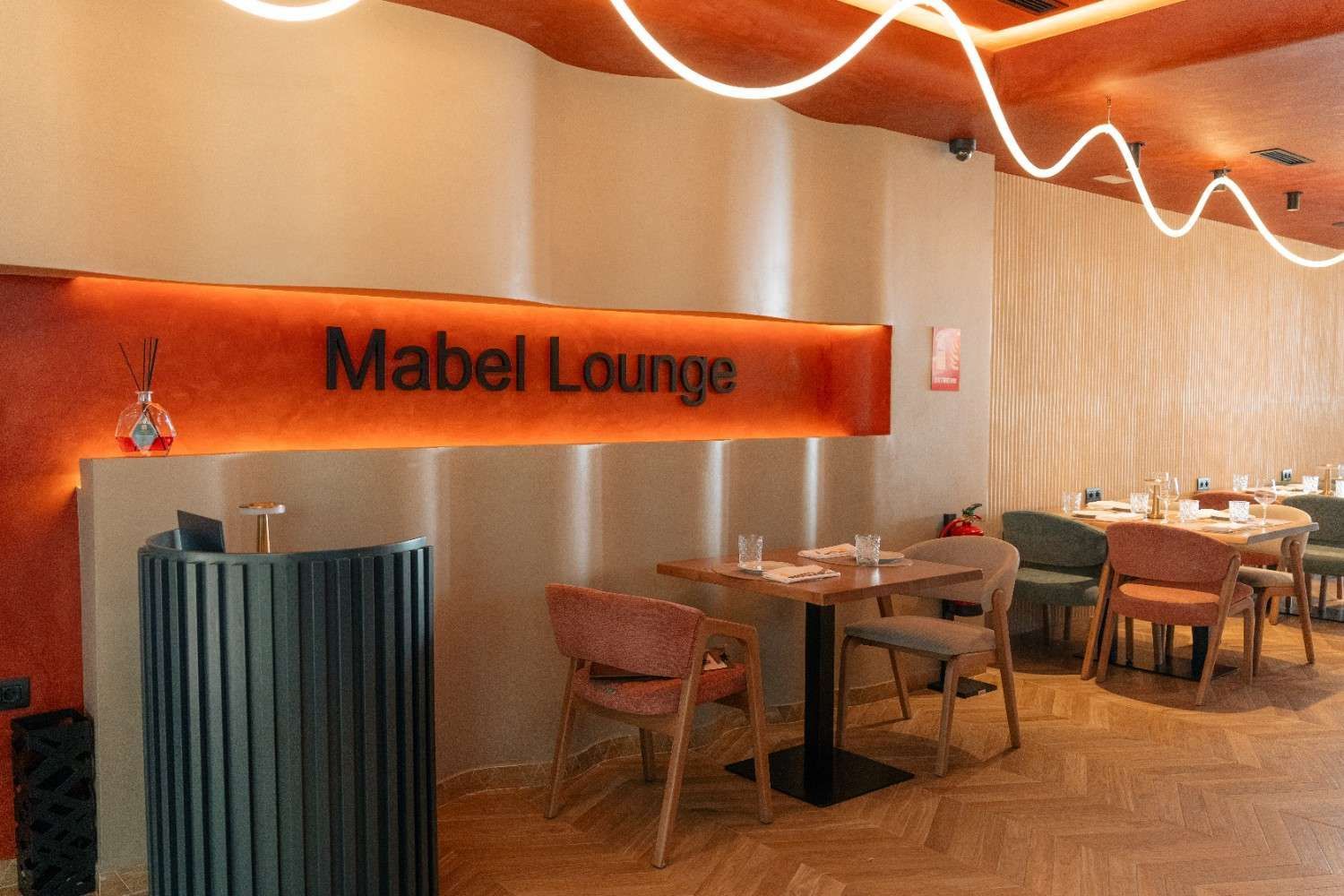 Entrada al Mabel Lounge con letrero naranja, mesas, sillas y luces cálidas en el techo dentro de un área de comedor.