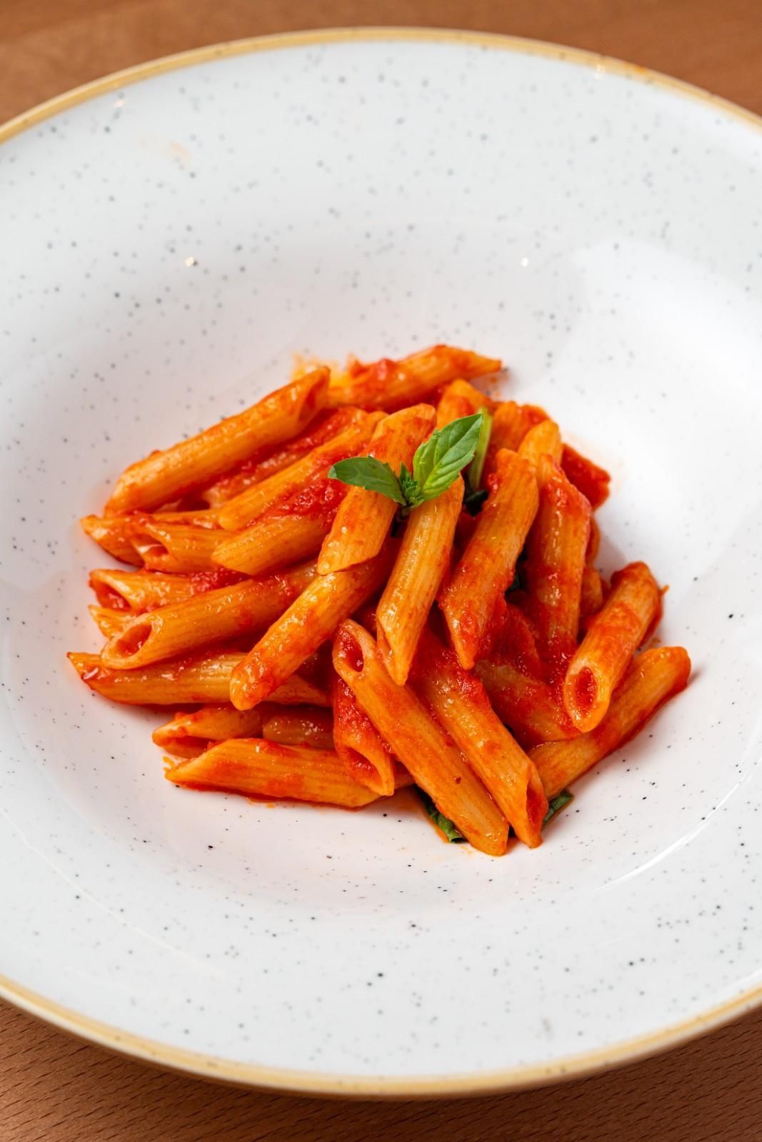 Plato de pasta penne en salsa de tomate roja con guarnición de albahaca sobre un plato blanco.