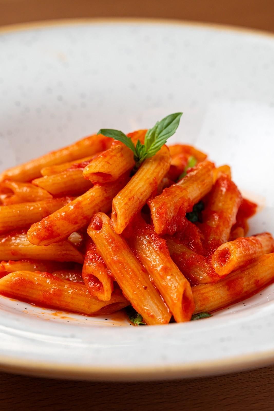 Pasta penne en salsa de tomate rojo, cubierta con albahaca, servida en un tazón blanco.