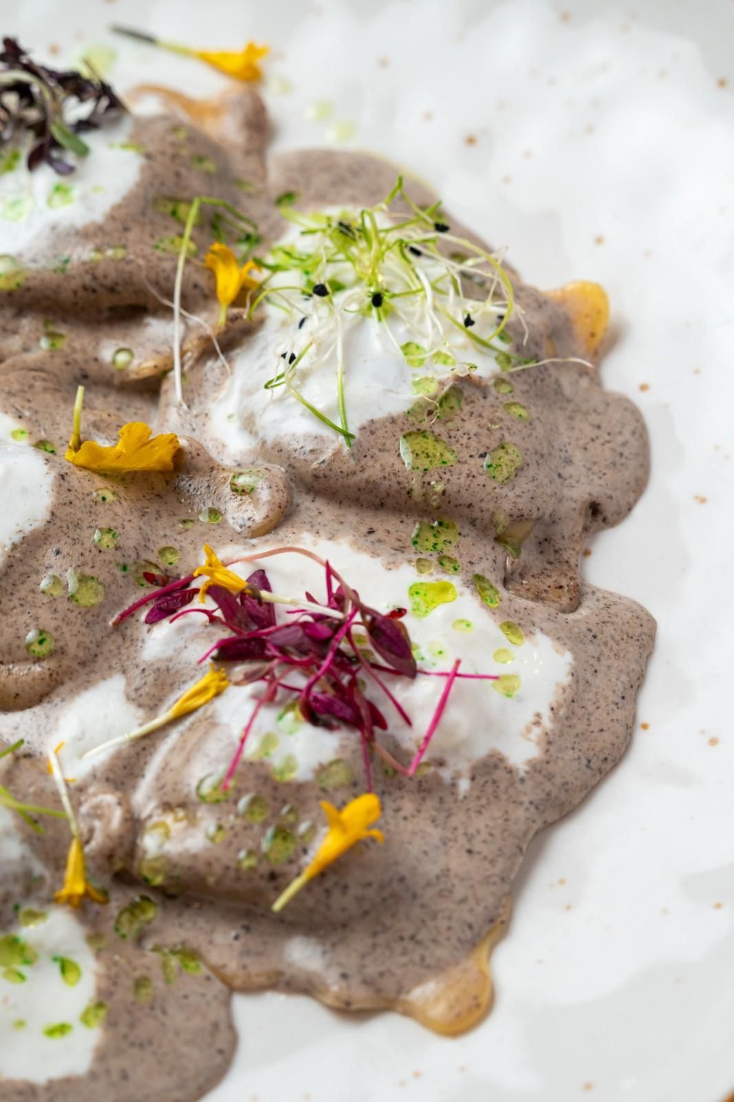 Plato cremoso gris con salsa blanca, hierbas verdes y flores comestibles amarillas y moradas sobre un plato blanco.