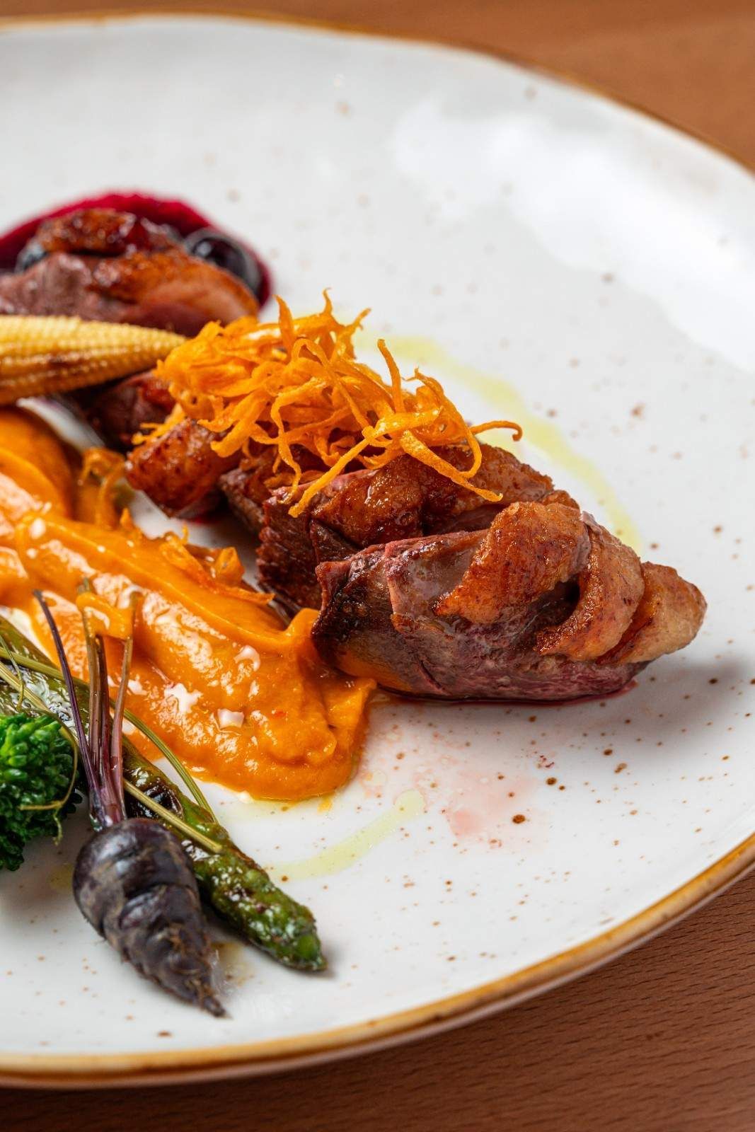 Filete de pescado gourmet emplatado con salsa de naranja, espárragos y guarnición desmenuzada en un plato blanco.