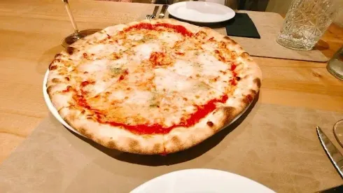 Una pizza de queso sobre una mesa de madera con platos y un vaso cerca.