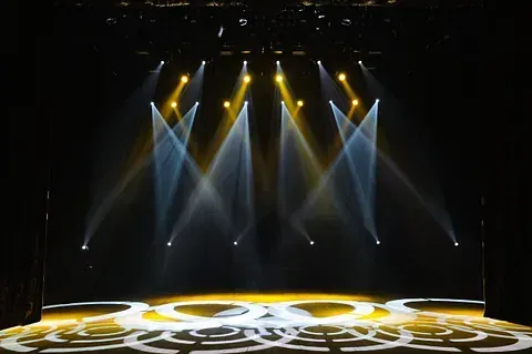 Escenario iluminado con focos amarillos y blancos, que proyectan patrones en el suelo, sobre un fondo oscuro.