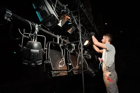 Un hombre con mono ajusta las luces del escenario suspendidas de una estructura de metal en un teatro oscuro.