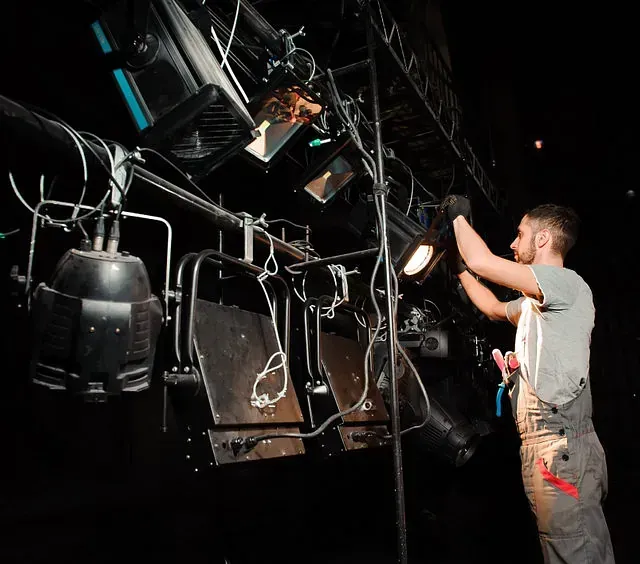 Una persona ajusta un foco en un escenario. Lleva un mono y trabaja en la iluminación superior en un teatro oscuro.