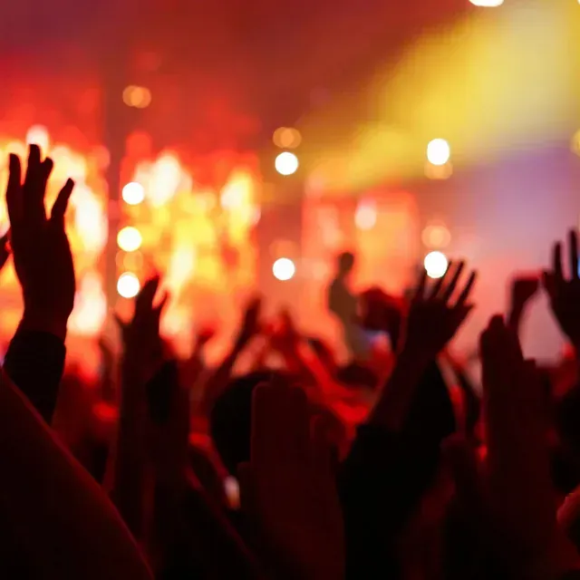 Manos levantadas entre la multitud en un concierto, bañadas por luces rojas y amarillas del escenario.