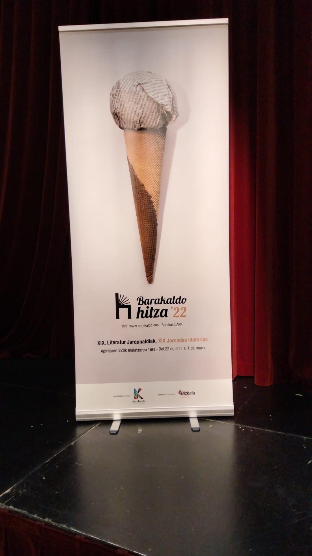 Una pancarta con la ilustración de un cono de helado. La pancarta está en un soporte sobre un escenario, con un fondo oscuro.