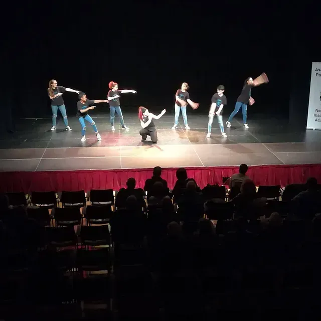 Una actuación de danza en un escenario con artistas vestidos con camisas negras y vaqueros, frente a un público en un teatro.