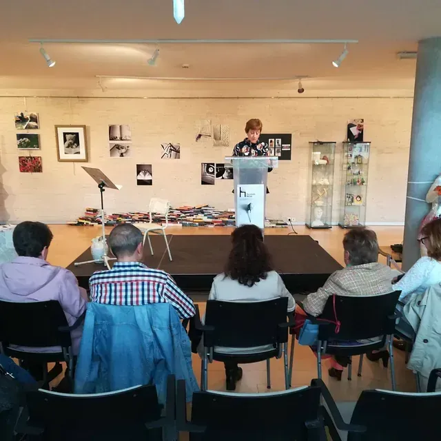Una mujer lee desde un podio en un pequeño escenario a una audiencia en una sala con arte en las paredes.