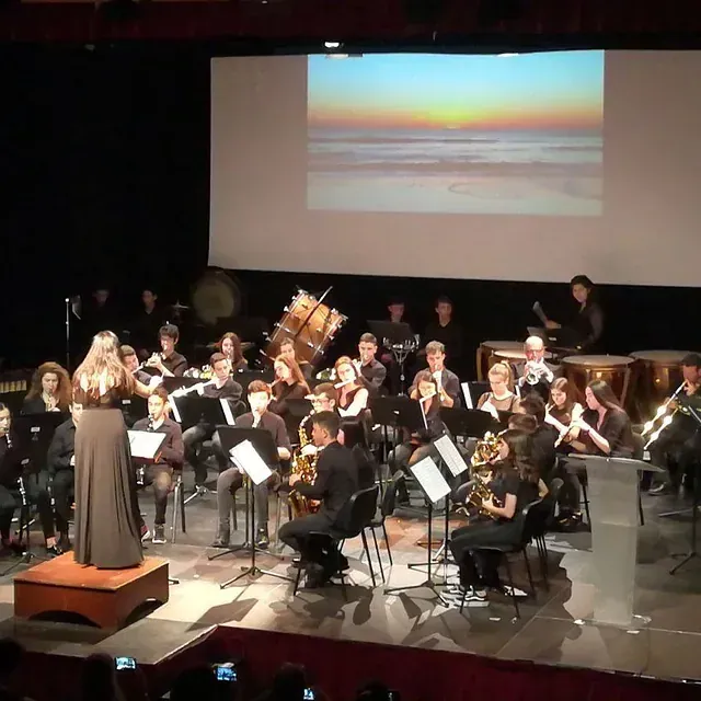 Un coro y una orquesta actúan en el escenario. Una mujer con vestido canta, con músicos detrás y una puesta de sol en la playa en la pantalla.