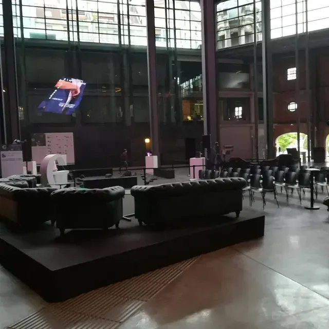 Vista interior de un espacio grande y moderno para eventos con sofás negros en una plataforma, filas de sillas y una pantalla grande que muestra a una persona.