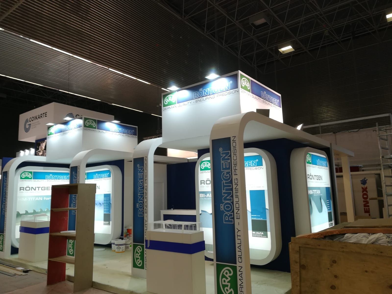 Un stand en una feria comercial; señalización azul, blanca y verde. Elementos arquitectónicos curvos frente a un techo oscuro.