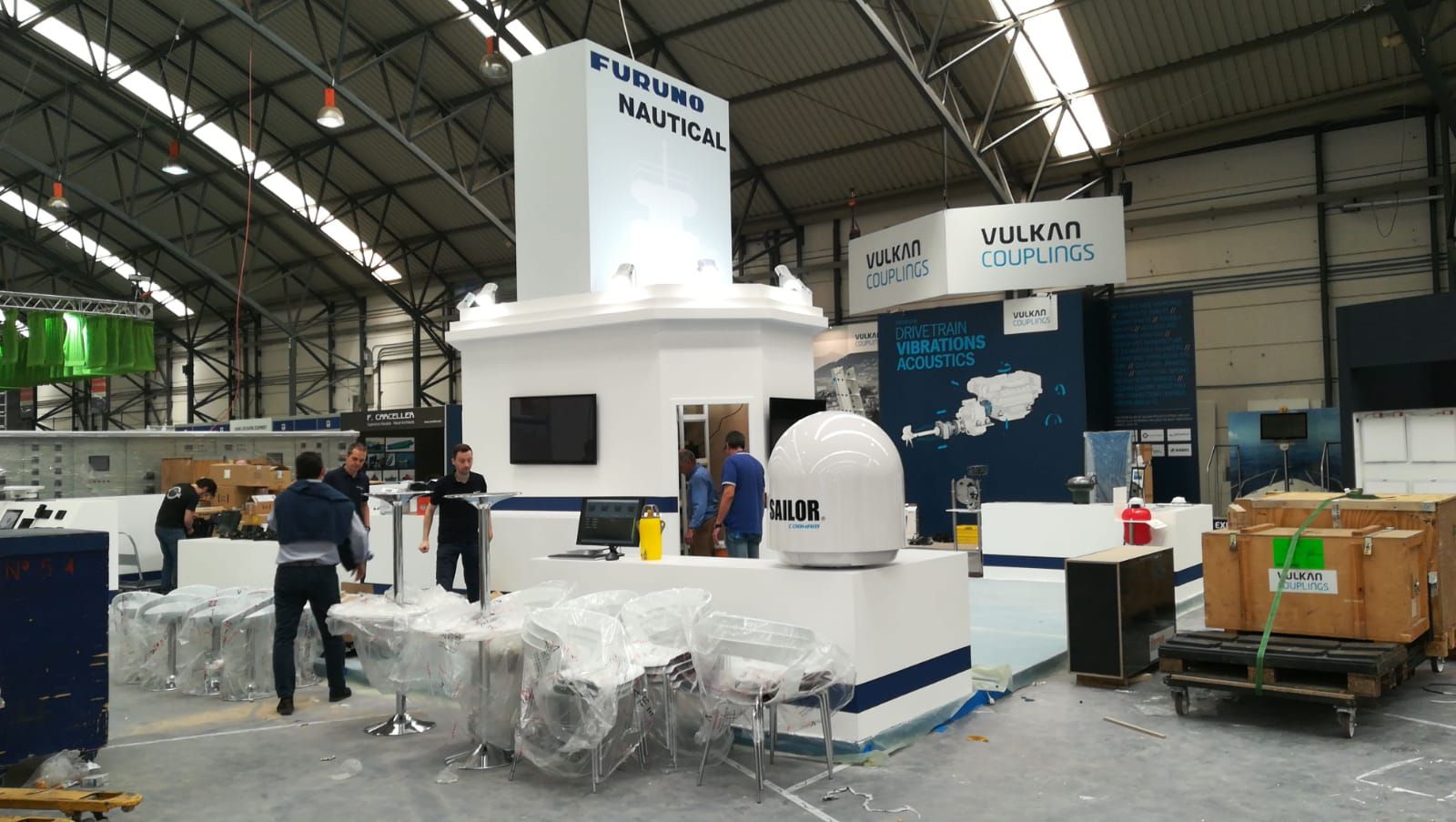 Un stand de exhibición interior, probablemente de una empresa de electrónica marina. Presenta estructuras blancas con detalles y logotipos azules, y algunas personas trabajando a su alrededor.