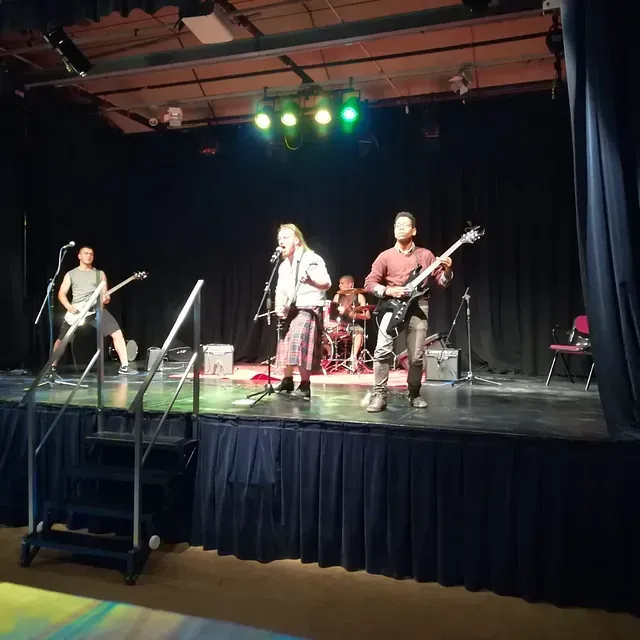 Banda tocando en un escenario con un cantante, dos guitarristas y un baterista. El escenario está iluminado con luces verdes y amarillas sobre un fondo negro.