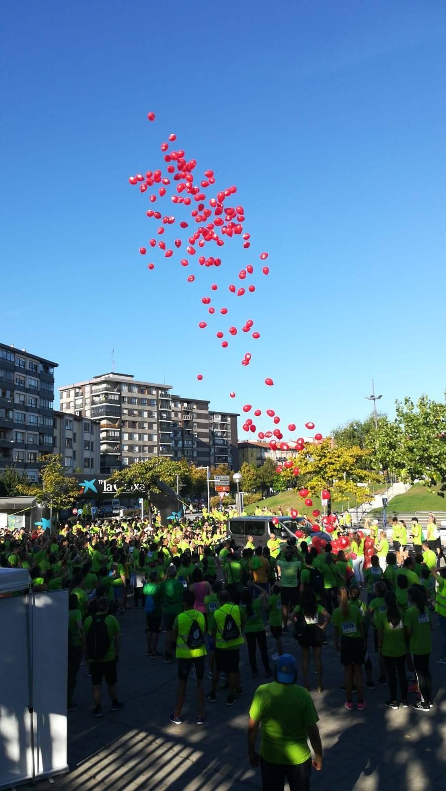 Una multitud de personas con camisetas verdes y chalecos amarillos lanza un grupo de globos rojos a un cielo azul claro.