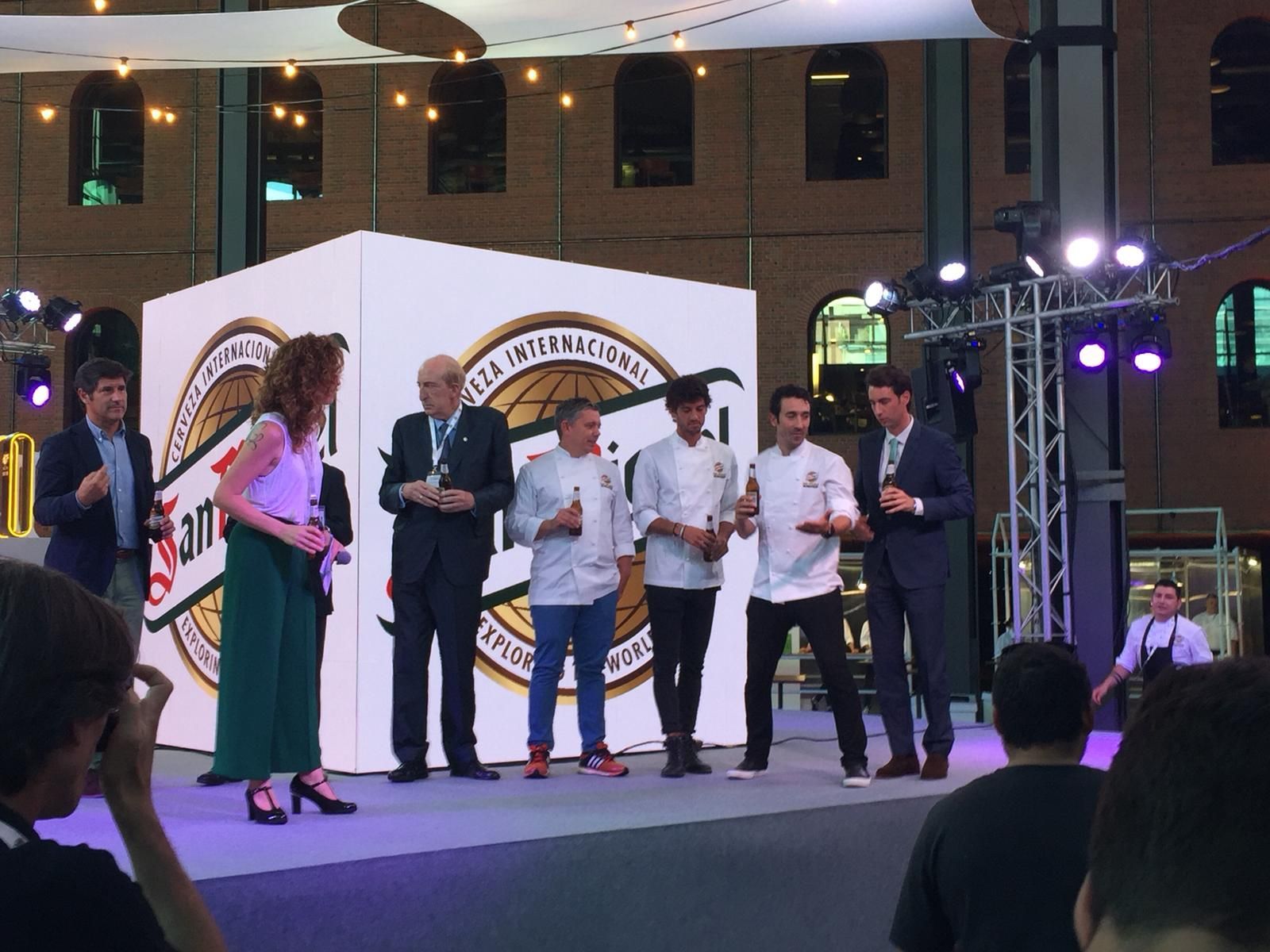 Personas en el escenario, algunas con trofeos, en una ceremonia de premios. Un gran logotipo se ve detrás de ellas, frente a un edificio con ventanas arqueadas.