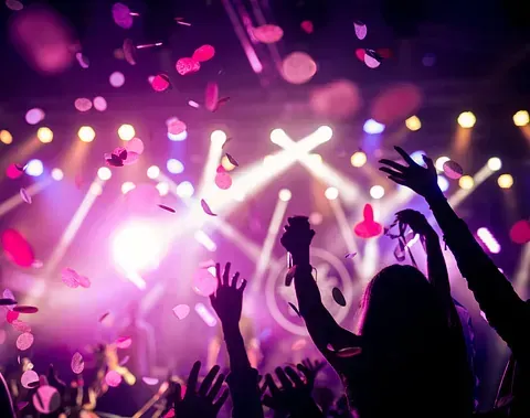 Personas con los brazos en alto en un concierto. Cae confeti contra las luces del escenario; se ven siluetas al frente.