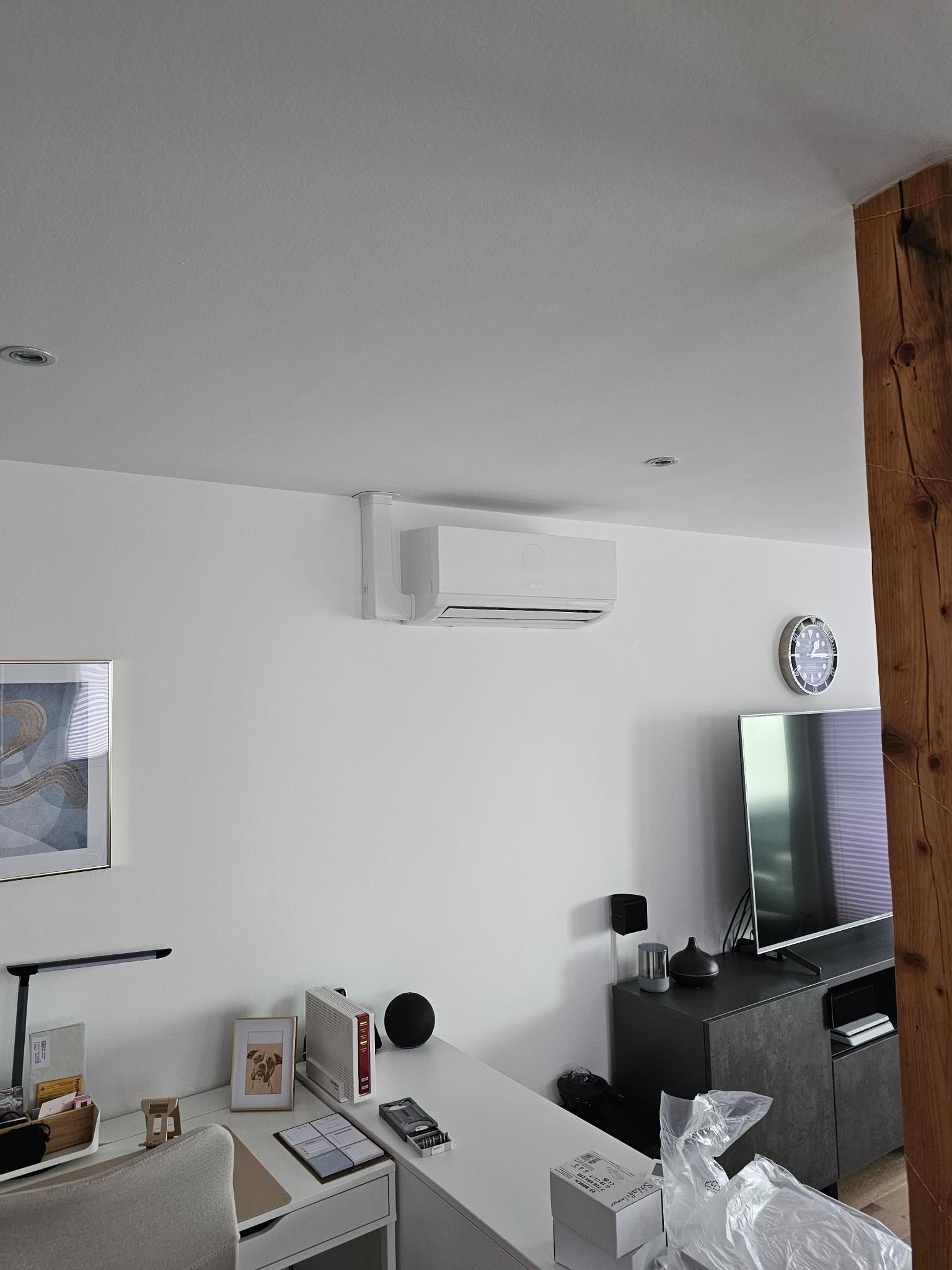 Modernes Wohnzimmer mit weißen Wänden, Fernseher, Uhr, wandmontierter Klimaanlage und einem Schreibtisch im Vordergrund
