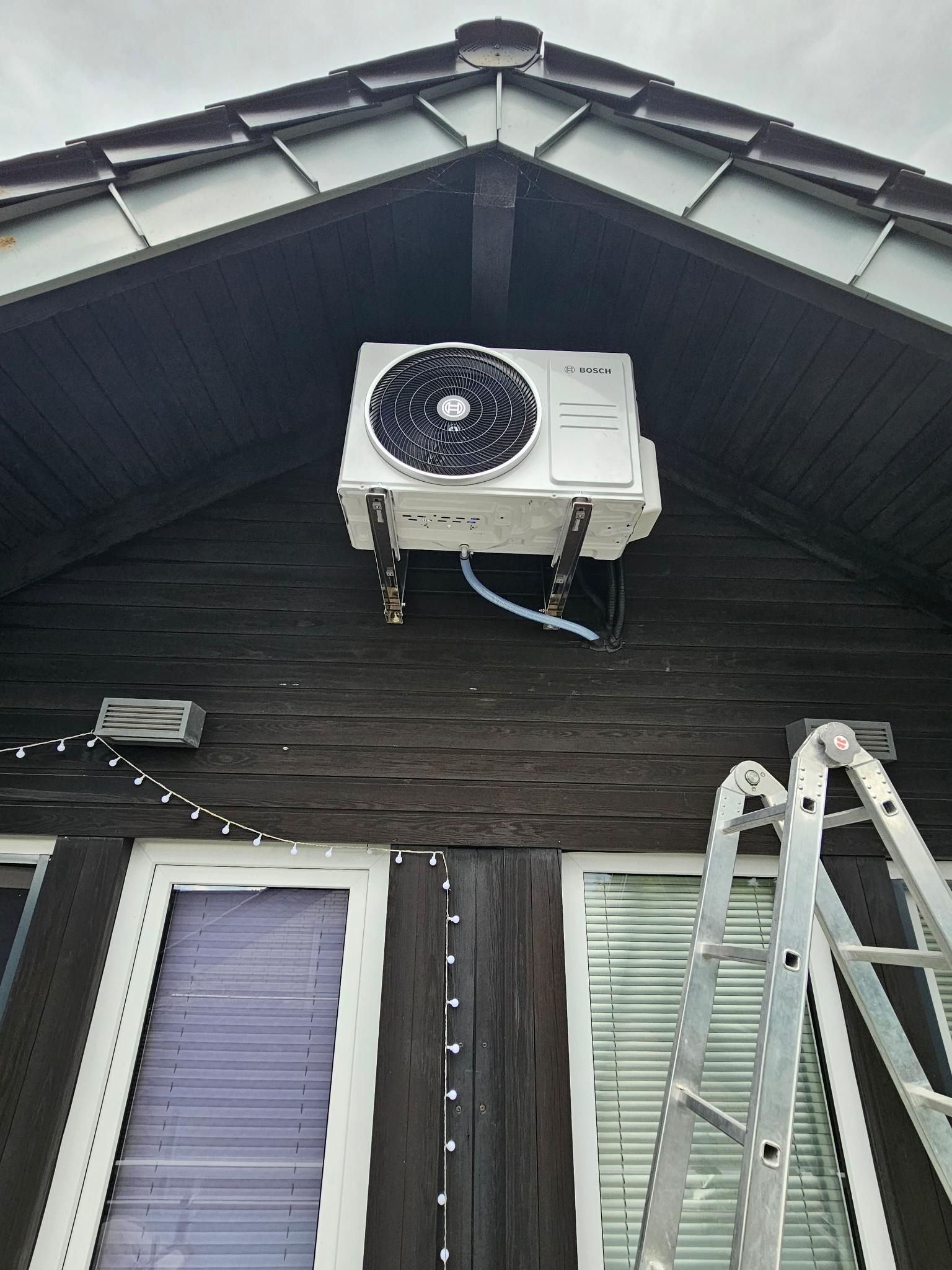 Weiße Außenklimaanlage, hoch oben an der dunklen Hausfassade montiert, mit einer Leiter am rechten Rand.