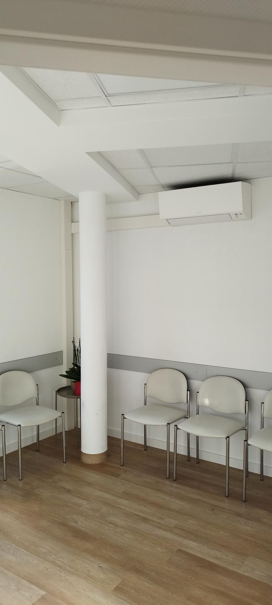 Kleiner Wartebereich mit weißen Stühlen, einer Säule, Holzboden und wandmontierter Klimaanlage.