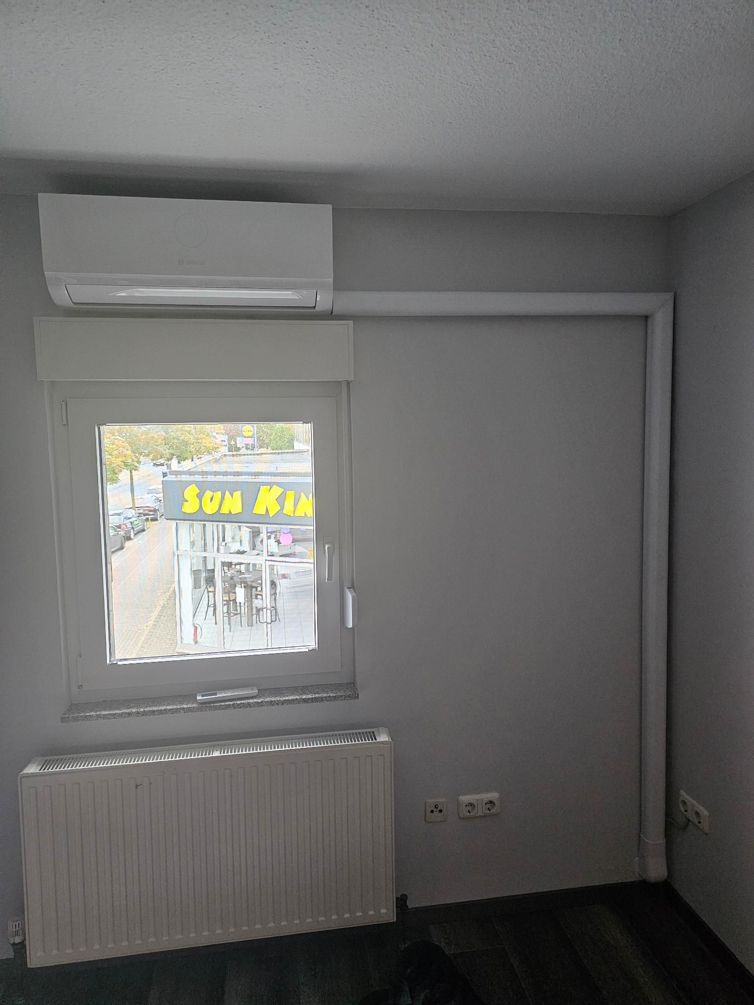 Leeres Zimmer mit einer an der Wand montierten Klimaanlage, einem Fenster und einem Heizkörper neben einer geschlossenen Tür.