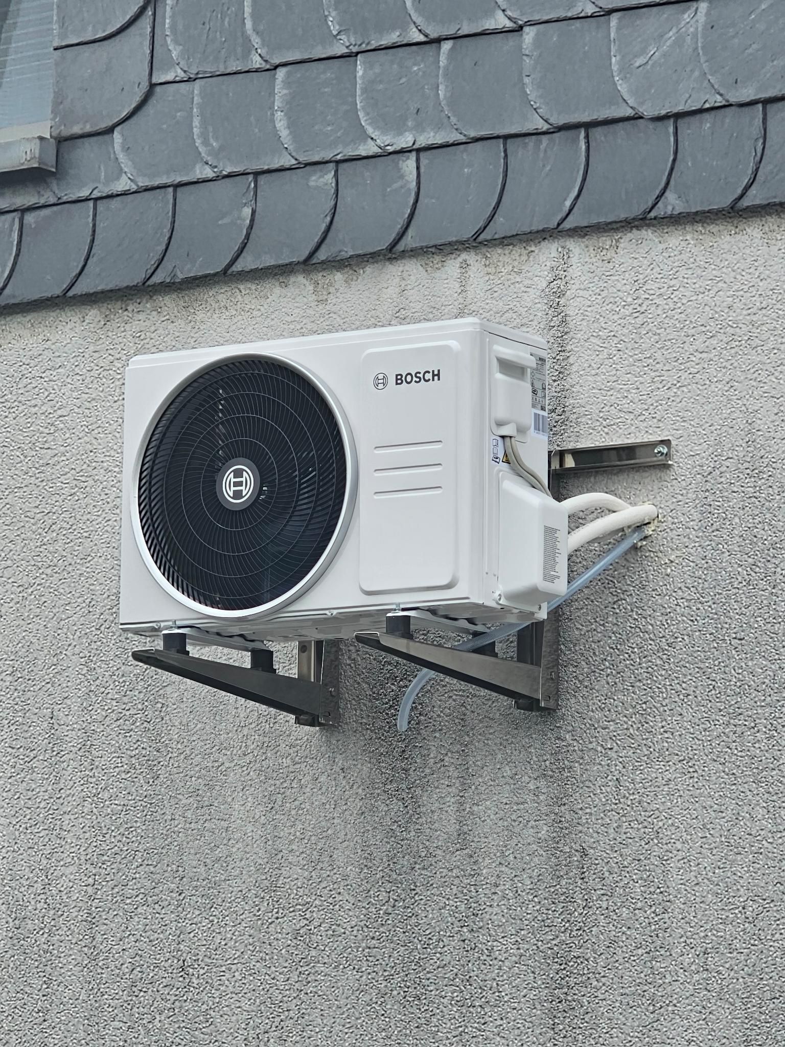 Außenwandmontierte Klimaanlage an einem verputzten Haus unter grauen Schindeln.