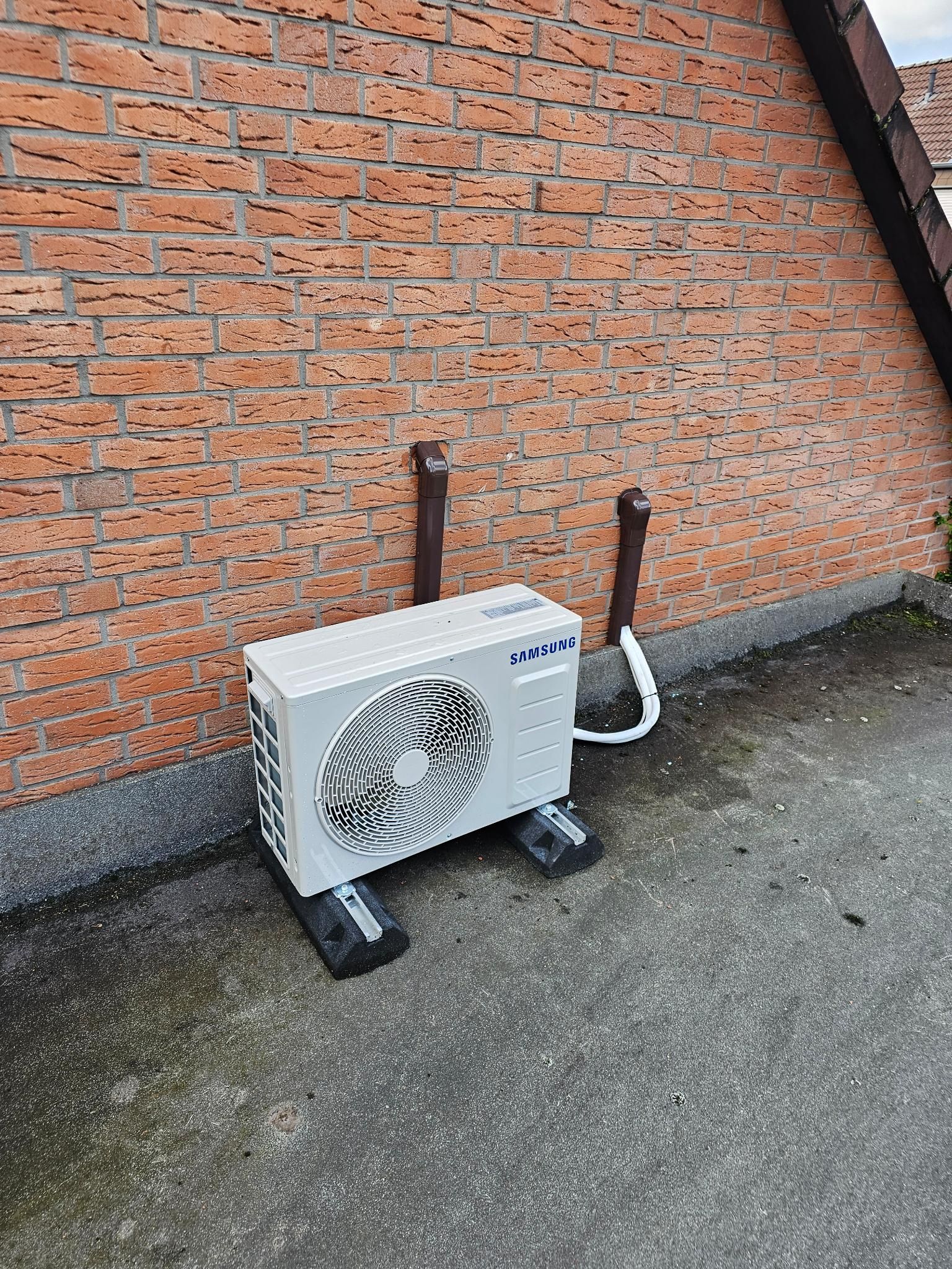Außengerät einer Klimaanlage auf Beton neben einer Ziegelmauer mit zwei Rohren.