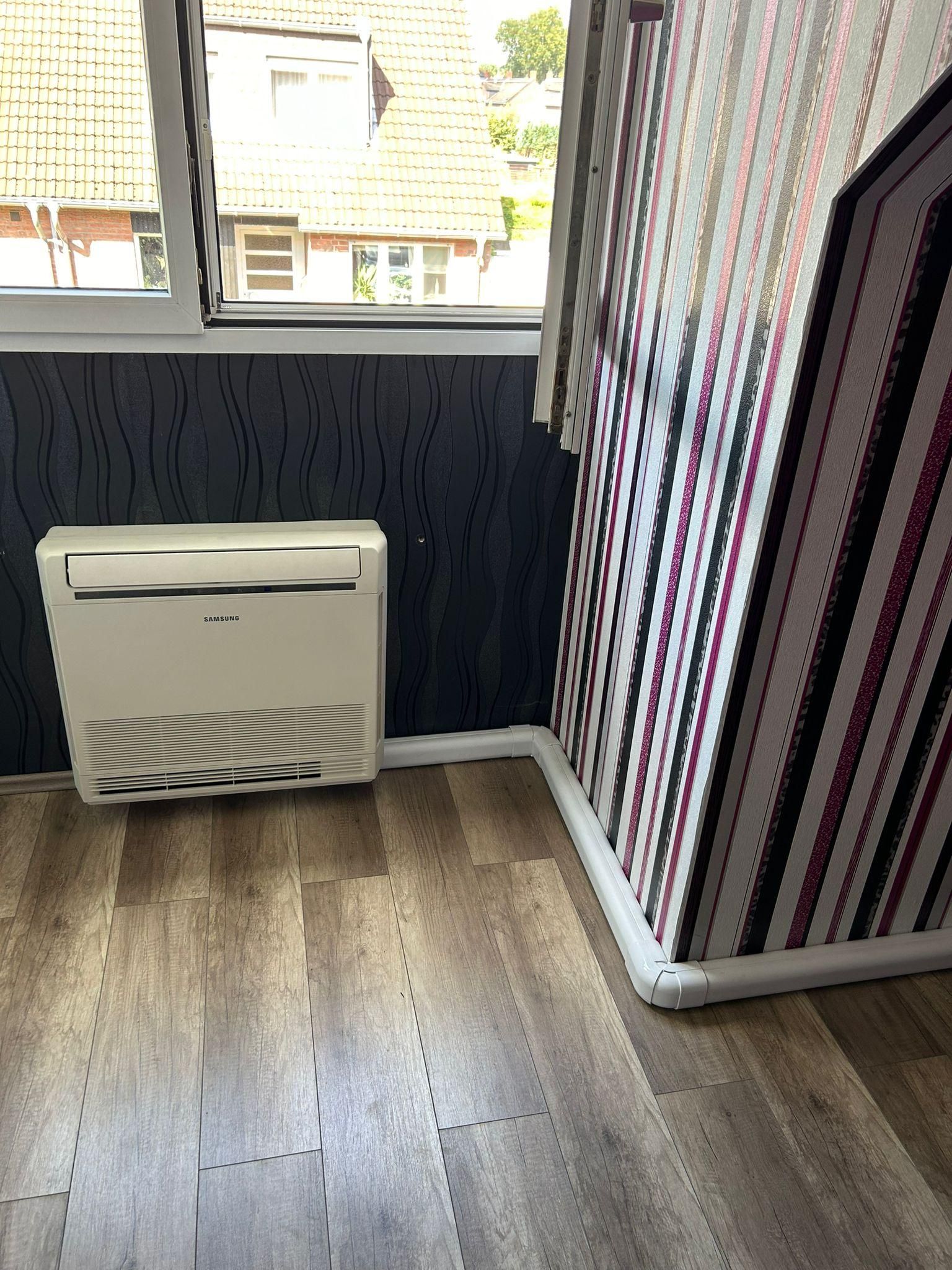Kleines Zimmer mit gestreifter Tapete, Holzboden und einer an der Wand montierten Klimaanlage unter dem Fenster.