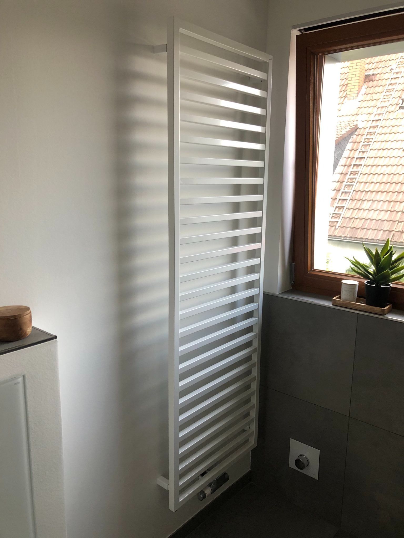 Ein weißer Wandheizkörper neben einem Fenster mit Jalousien und einer Topfpflanze auf der Fensterbank.