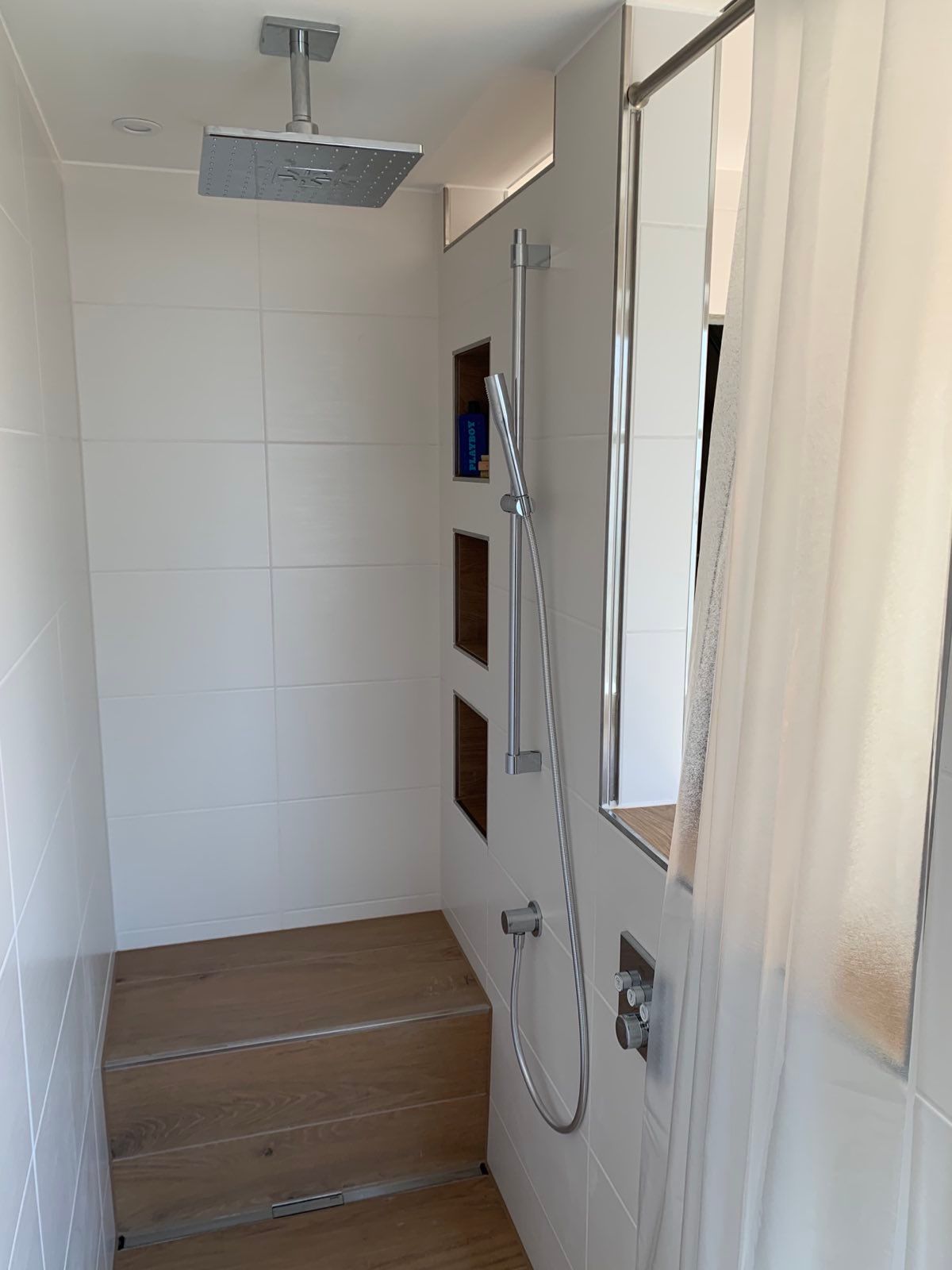 Kleines Badezimmer mit Dusche, weiß gefliesten Wänden, Holzboden und einem Fenster mit Vorhang.