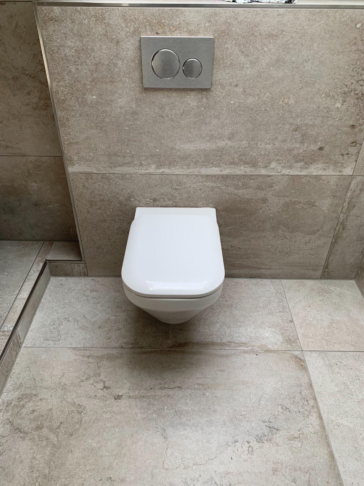 Kleine, weiße, wandhängende Toilette in einem gefliesten Badezimmer, darüber befindet sich ein Zweimengenspülknopf.