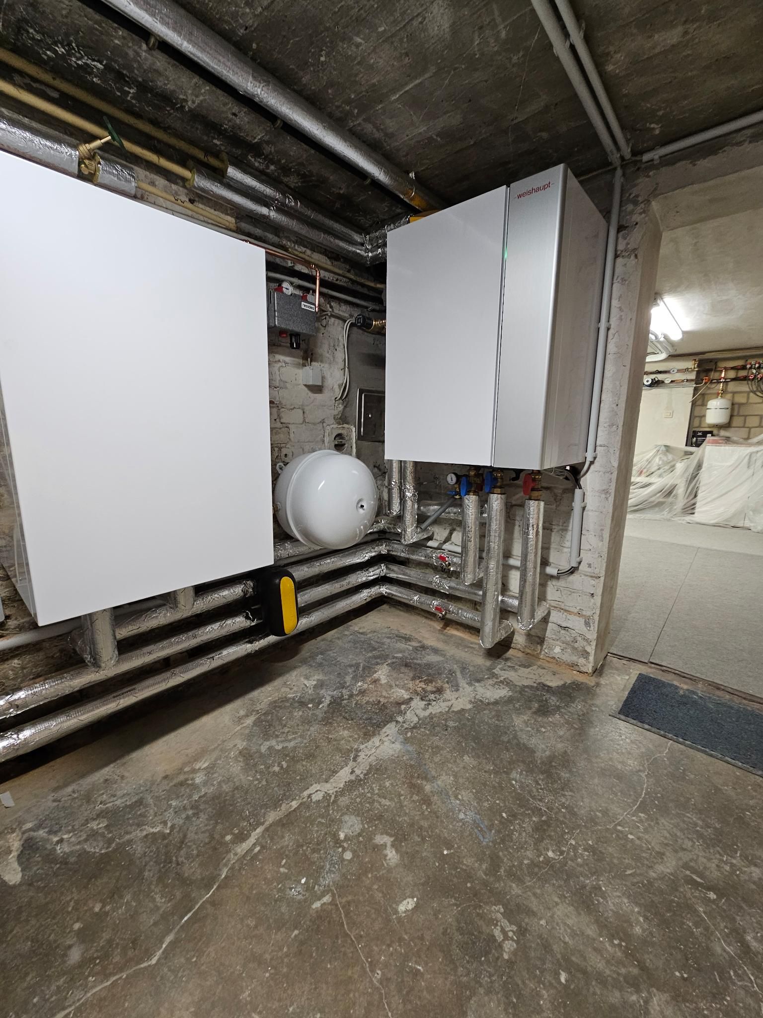 Hauswirtschaftsraum im Keller mit weißen Elektroschränken, Rohren und einem kugelförmigen Tank an Betonwänden.