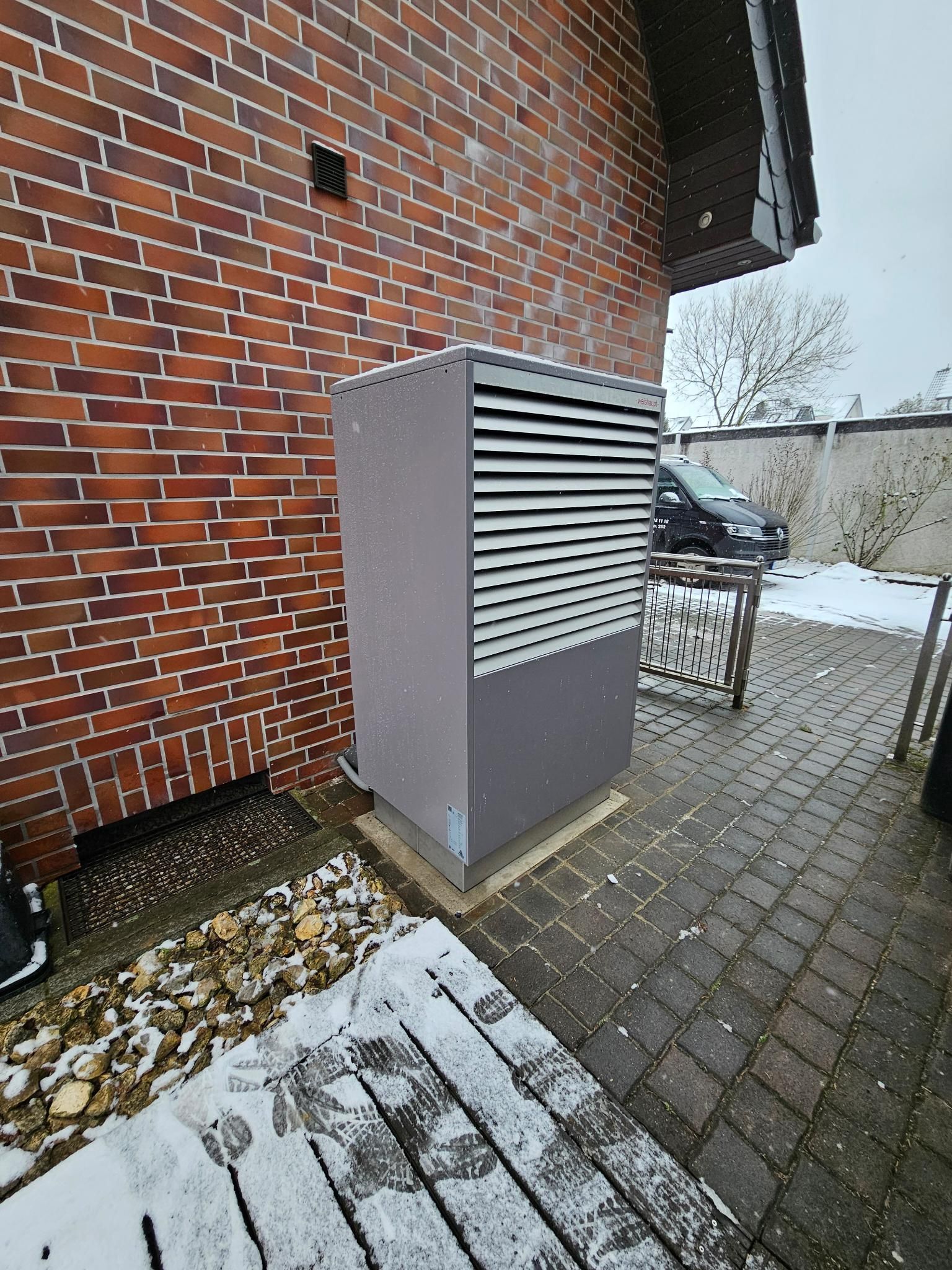 Eine graue Wärmepumpe neben einer roten Backsteinmauer auf einer schneebedeckten, gepflasterten Terrasse