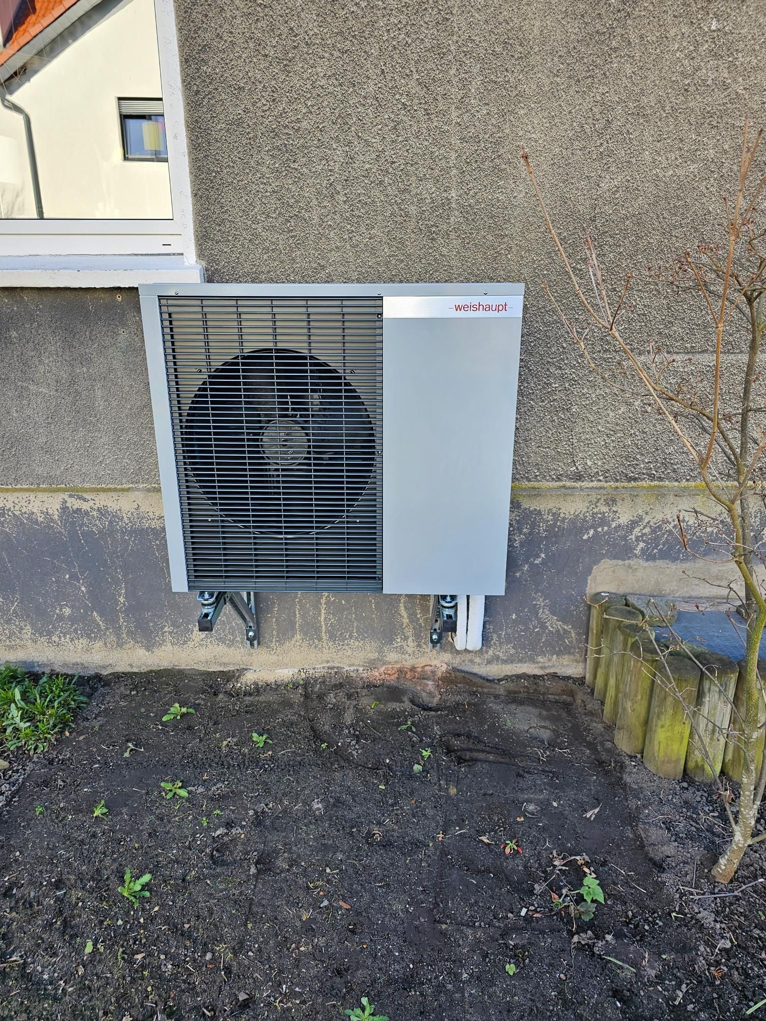 Graue Außeneinheit der Klimaanlage neben einer Gebäudewand, hinter deren Gitter ein Ventilator sichtbar ist.